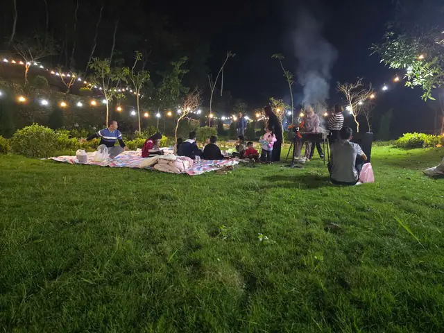 Bãi cỏ buổi tối giăng dây đèn, khách ngồi nướng BBQ, chụp từ xa