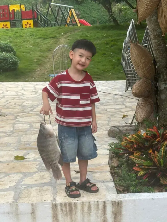 Một bé trai áo sọc đỏ cầm con cá trên lối đá, phía sau là sân chơi có cầu trượt
