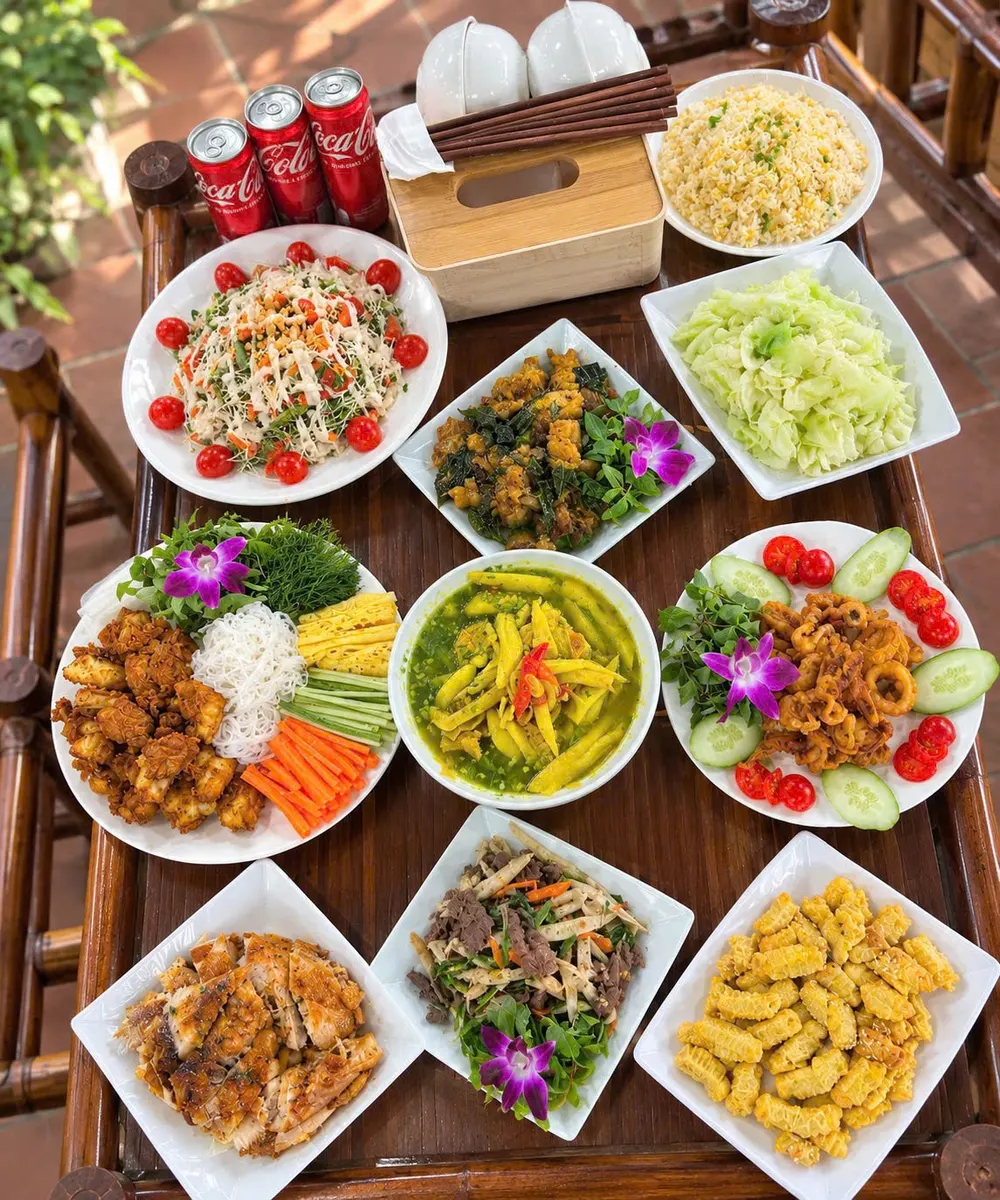 Mâm cơm quê nhìn từ trên xuống: nộm, cá, gà, rau xào, canh và đĩa chả bày trên bàn gỗ