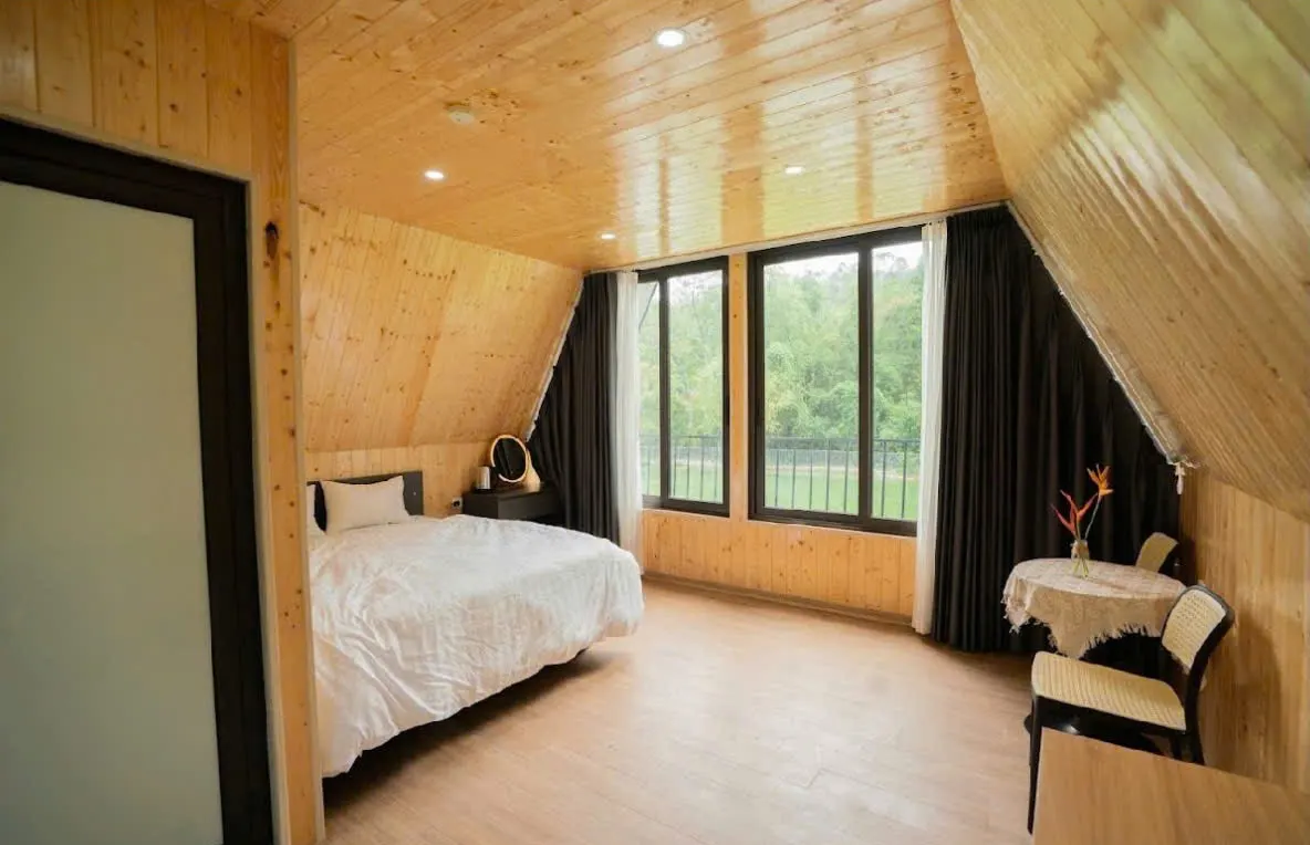 Bên trong bungalow mái chữ A: giường đôi, vách gỗ sáng màu, cửa sổ kính nhìn ra cây xanh