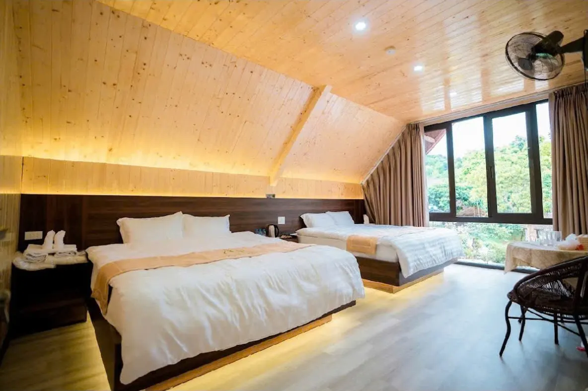 Phòng gia đình trần gỗ thông với hai giường đôi trải ga trắng, cửa kính lớn nhìn ra vườn cây
