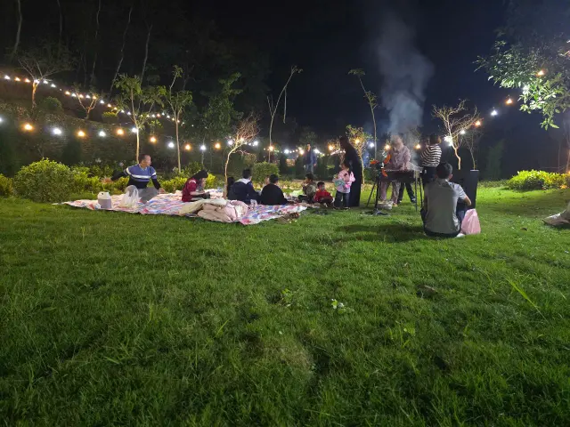 Nhóm khách ngồi picnic trên bãi cỏ buổi tối, dây đèn vàng giăng quanh và khói BBQ bên xa